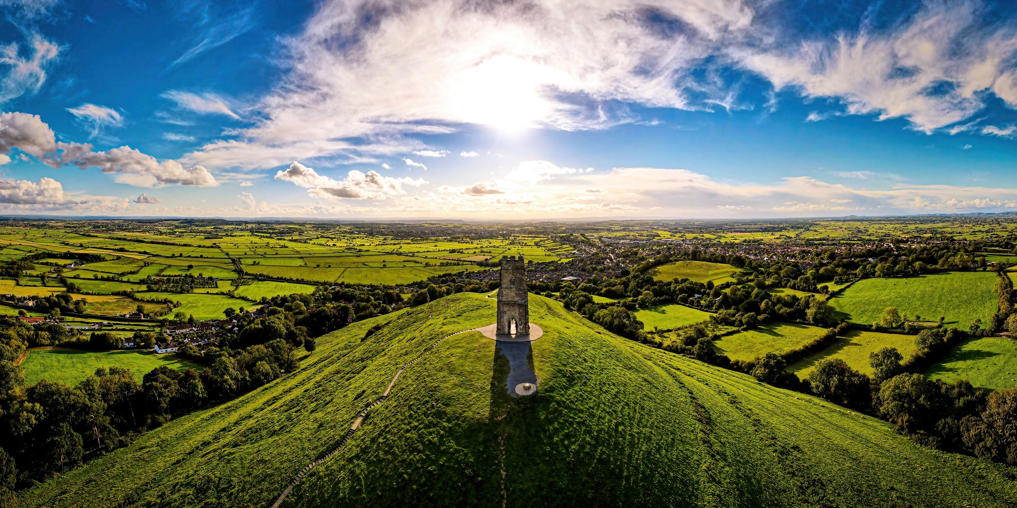 A drone shot of Glastonbury Tor, from above. The view of the green countryside appears to go on for miles, set against a vivid blue sky